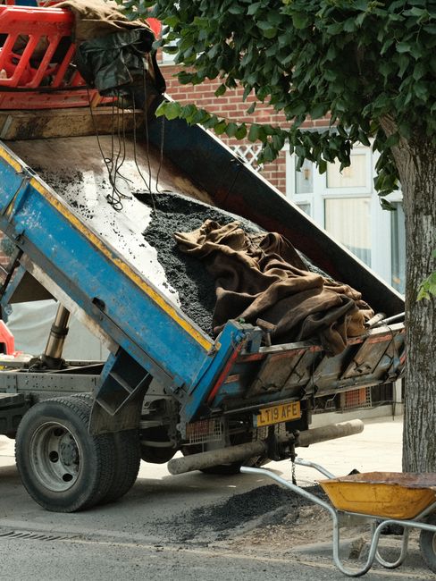A small blue dump truck is parked on the street with its bed tilted upwards, unloading a pile of dark asphalt or black gravel onto the ground. The truck's tailgate is open, revealing various tools, including a brown cloth and other packing materials, situated on the load. The truck's rear registration plate reads 'L119 AFE.' Behind the vehicle, there is a brick building with a window, and a leafy tree with green foliage partially obscures the scene. The environment suggests a home improvement or driveway resurfacing project, with a yellow wheelbarrow positioned nearby, indicating ongoing construction or landscaping work. This image relates to home relocation or property maintenance tasks, which are often coordinated by businesses like Man with Van Whitton for efficient house removals and moving services.