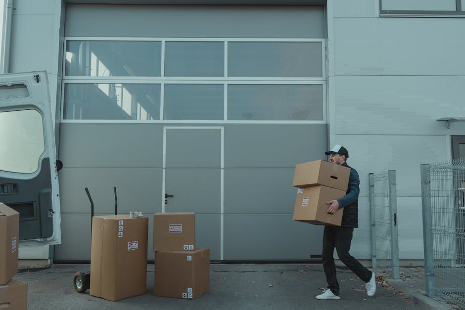 A man wearing a black baseball cap, dark jacket, and jeans is outside a modern industrial building with large glass window panels and grey metal walls. He is carrying two medium-sized cardboard boxes,