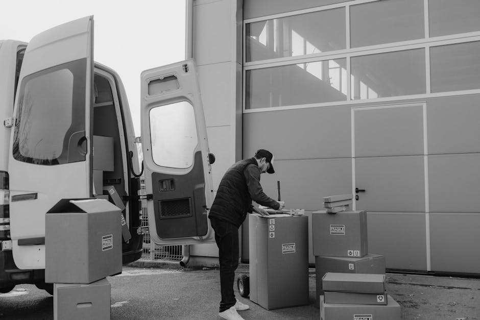 A man wearing a black baseball cap, dark jacket, and jeans is outside a modern industrial building with large glass window panels and grey metal walls. He is carrying two medium-sized cardboard boxes,