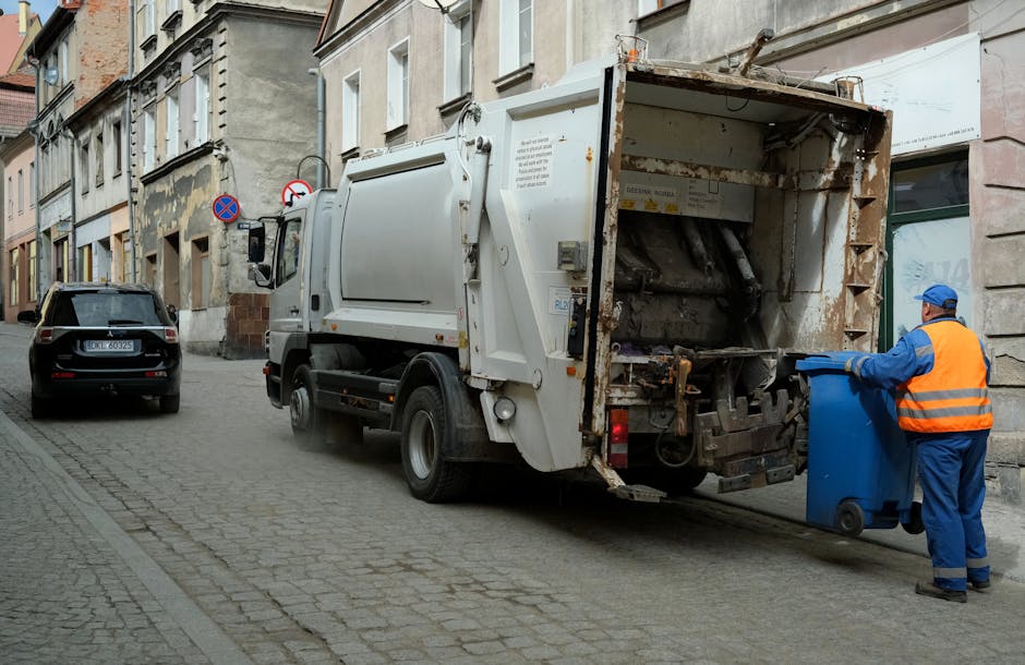 A sanitation worker wearing a blue uniform and an orange safety vest is seen loading a blue wheeled bin into a white garbage collection truck parked on a cobblestone street. The truck's rear compartment is open, revealing rusted steel interior and hydraulic lifting mechanisms used in waste collection. The street is lined with older, multi-storey residential buildings with weathered facades and various signs indicating parking restrictions. A black car is parked adjacent to the waste truck, and there is soft natural lighting suggesting daytime. The scene depicts part of a home relocation or clearing process, with the worker handling waste materials as part of general rubbish collection services, with Man with Van Whitton providing such removals or disposal support in the Whitton (TW2) area.