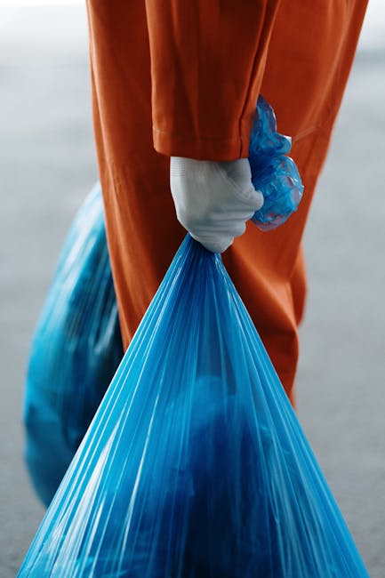 A person wearing an orange protective jumpsuit and white gloves is holding a large blue rubbish bag filled with waste, standing outdoors on a paved surface. In the background, a portion of a blue plastic waste bag is visible on the ground. The scene suggests waste collection or disposal, which may be part of a home relocation or clearance process. This image aligns with services provided by Man with Van Whitton, such as removals, packing, and waste removal, supporting efficient moving logistics and home clearance in Whitton (TW2). The lighting is natural, highlighting the focus on waste handling in a professional context related to furniture transport or property clearance.