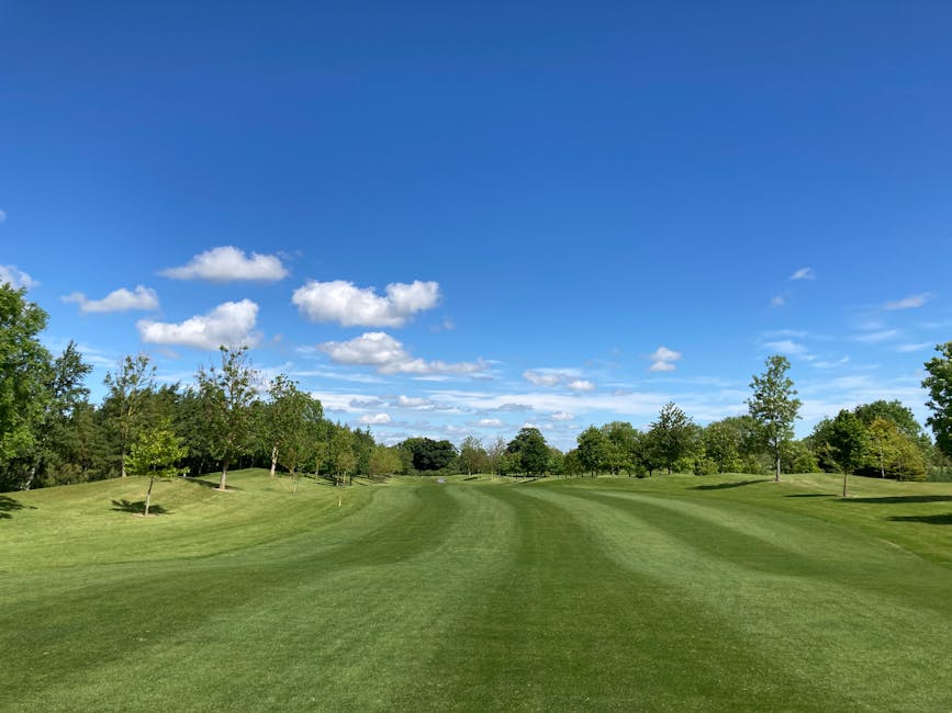 A wide view of a well-maintained green golf course with evenly mowed grass and a gentle slope, bordered by a mix of deciduous trees with lush green foliage. The sky is clear with a bright blue hue and scattered cumulus clouds. In the foreground, the grass is distinctly striped, indicating recent mowing. The landscape extends into the distance with no visible human activity or structures. The scene provides an open, natural environment, suitable for recreational activities like golf or outdoor relaxation. This tranquil setting captures the outdoor space surrounding an area where house removals or home relocation services, such as those offered by Man with Van Whitton, could involve logistical planning for accessing properties with varied surrounding environments.