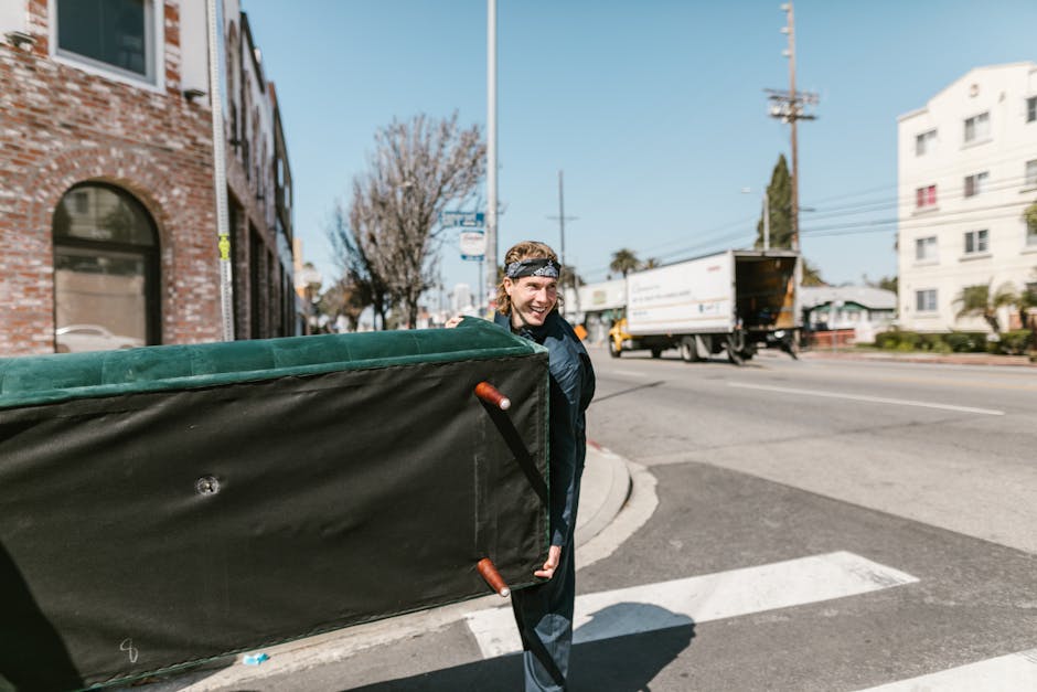 A man with shoulder-length hair wearing a headband and a dark jacket is standing outdoors on a street, carrying a large padded furniture protectors or mattress cover with both hands. Behind him, he is positioned near a curb at the corner of a residential area with multi-storey brick buildings, trees, and utility poles visible. A moving van or truck is parked nearby, partially loaded, with the back open and furniture or boxes possibly visible inside. The street is busy with vehicles, including another truck in motion in the background, and a zebra pedestrian crossing is visible in the foreground. The scene is illuminated by natural daylight, suggesting a clear sky. This image reflects aspects of home relocation and furniture transport processes, with [COMPANY_NAME], Man with Van Whitton, providing removals and moving services, as part of a professional packing and loading operation used in house moves to Whitton High Street.