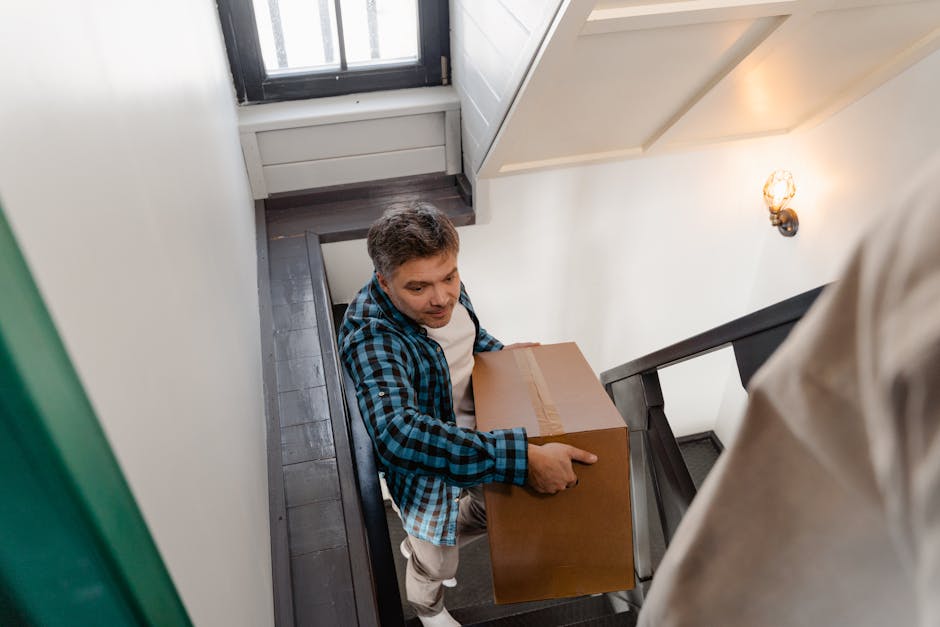 A man wearing a blue and black checkered shirt is carrying a medium-sized cardboard box on a staircase inside a house during a home relocation. The staircase features dark wooden steps, and the man is holding the box steadily with both hands, preparing to descend or ascend. The interior walls are painted white, and a black handrail runs along the staircase. Natural light filters in from a small window at the top of the stairs, illuminating the scene. A small wall-mounted light fixture with a decorative bulb is visible on the right side. This action is part of the furniture transport process involved in packing and moving, demonstrating careful handling during house removals. The image highlights the logistical process carried out by professional moving services like Man with Van Whitton, ensuring safe and efficient furniture transport within the home environment.