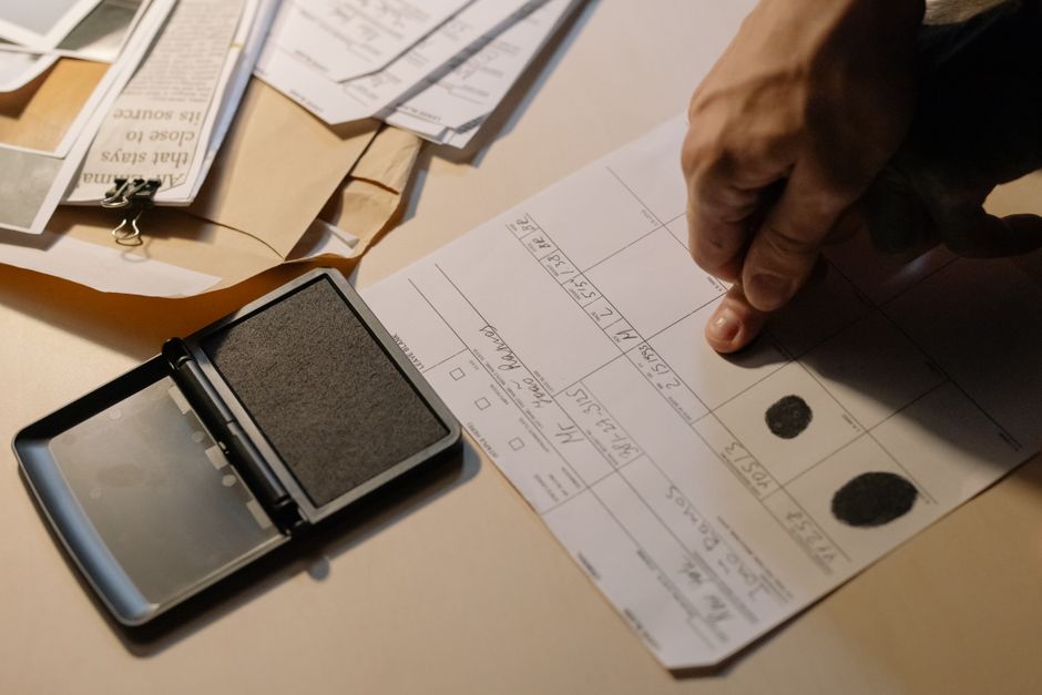 A close-up of a hand stamping a document with fingerprint ink, which is part of a legal or official paperwork process related to house removals or probate clearances. The document is on a light-colored surface along with various other papers, some secured with clips, and a smartphone with an attached stamp device is visible nearby. The setting appears to be inside a property, possibly during the packing or legal verification stages of a home relocation process. The background includes more papers and packaging materials, indicating a detailed administrative or legal step involved in the moving or clearance service offered by Man with Van Whitton. The lighting is soft and natural, highlighting the desk setup used in managing house removals or probate-related formalities.