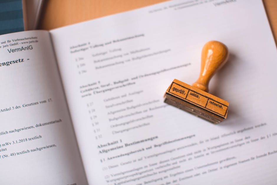 A close-up photograph showing an open legal document with printed text in German, laid out on a flat surface indoors. On top of the document is an orange rubber stamp with a wooden handle, positioned to the right side of the page. The background reveals a wooden table or desk, and the lighting is natural, casting soft shadows. This scene illustrates the process of handling legal paperwork relevant to probate clearance or estate management, which may be part of the home relocation or removal process undertaken by Man with Van Whitton when managing estate clearances and furniture transport during house removals in Whitton.