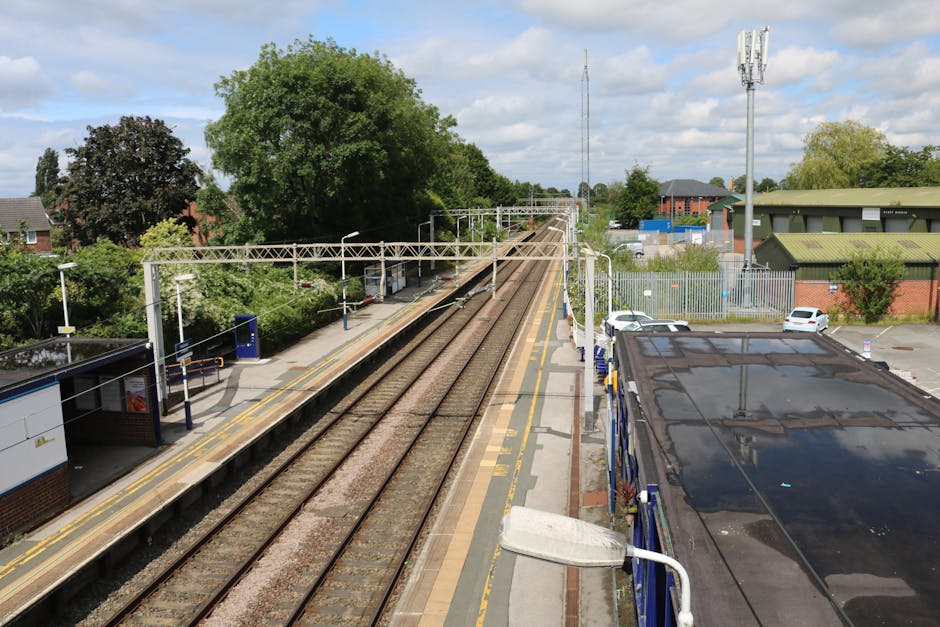 A view from a railway station platform shows multiple train tracks extending into the distance, with overhead electric lines supported by metal gantries. To the left, there is a small station shelter with a blue information kiosk and yellow tactile paving at the edge of the platform. To the right, a dark, reflective van with a slightly worn roof is parked adjacent to the platform, partially obscuring the view of a fenced area with several parked cars behind it. In the background, lush green trees, residential buildings, and communication masts can be seen under a partly cloudy sky. The scene depicts a typical urban train station environment, relevant to house removals or transport logistics, with the presence of vehicles and infrastructure supporting home relocation activities.