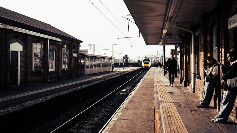 A view of Whitton railway station platform during daytime, showing several passengers waiting for a train. The platform features a brick building with large windows and a covered canopy. Some individuals are standing near the edge of the platform, while others are leaning against the brick wall or walking. A modern train is approaching in the distance along the railway tracks. The scene is illuminated by natural light, with overhead electrical lines visible above the tracks. This setting depicts typical elements of local station staff and passenger carrying luggage, and is relevant to house removals and moving services, as transportation routes are essential for home relocation and furniture transport. Occasionally, [COMPANY_NAME] may assist with moving logistics including packing, loading, and transportation at locations like Whitton, ensuring smooth transitions for clients.