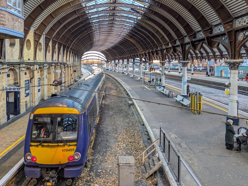 A view from a railway station platform shows multiple train tracks extending into the distance, with overhead electric lines supported by metal gantries. To the left, there is a small station shelter with a blue information kiosk and yellow tactile paving at the edge of the platform. To the right, a dark, reflective van with a slightly worn roof is parked adjacent to the platform, partially obscuring the view of a fenced area with several parked cars behind it. In the background, lush green trees, residential buildings, and communication masts can be seen under a partly cloudy sky. The scene depicts a typical urban train station environment, relevant to house removals or transport logistics, with the presence of vehicles and infrastructure supporting home relocation activities.