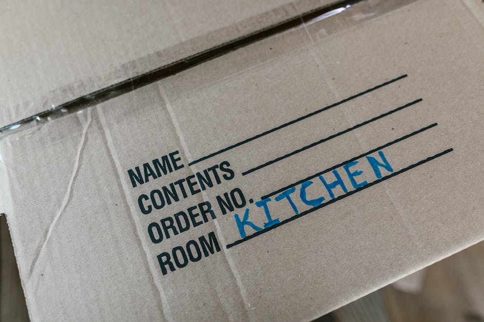 A close-up view of a cardboard moving box with printed labels, showing the handwritten word 'KITCHEN' in blue ink under the 'ROOM' label. The box appears to be part of a home relocation process, possibly prepared for a house move with [COMPANY_NAME], a professional removals service operating in Whitton. The box is sealed with clear packing tape along the top edge, and the surface surrounding the label is made of plain brown cardboard. The background includes a blurred view of other packed boxes and possibly a doorway or inside of a property, indicating an organized packing and moving environment. This image illustrates the packing and furniture transport aspects typical of house removals in connection with Richmond Council permits for Whitton relocations, highlighting the careful preparation involved in home relocation logistics.