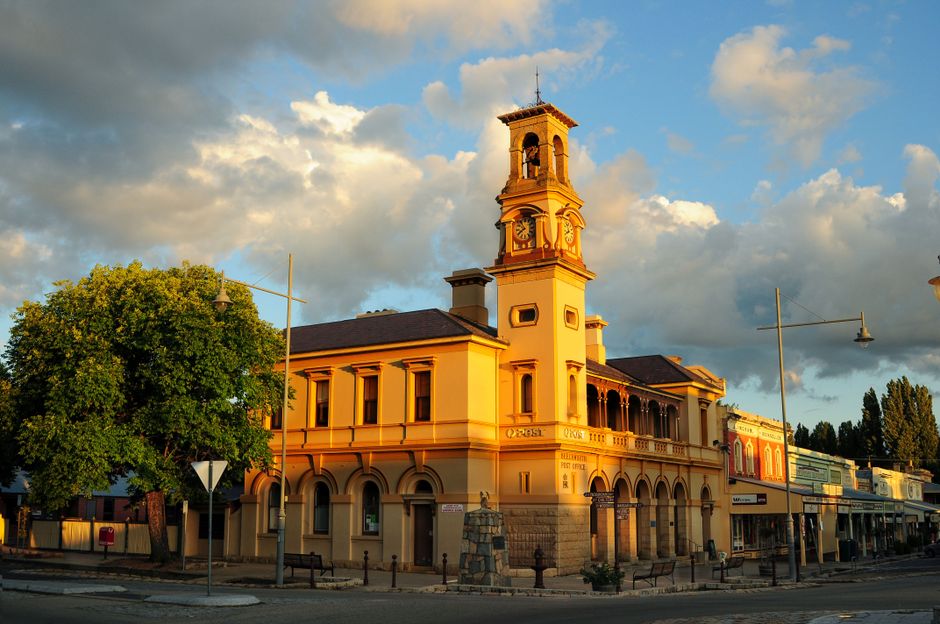 A historic building with a prominent clock tower and a decorative bell tower on top, situated on a street corner during late afternoon or early evening. The building features an arched ground floor with large windows, cream-colored walls with orange accents, and a dark roof. To the left, a green tree partially obscures the structure, while street signs and lamp posts are visible in the foreground. The sky above is partly cloudy, lit by warm, golden sunlight, creating long shadows on the street and highlighting the architectural details. In the vicinity, there are commercial premises and street signage, indicating an urban setting. This scene illustrates a typical location where house removals or furniture transport might consider the local environment and logistics, as seen in services provided by companies like Man with Van Whitton, which handles local removals and home relocation processes.