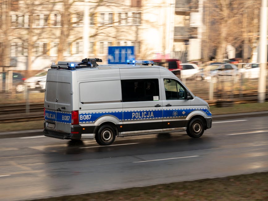 A silver police van with blue and white markings, displaying the word 'Policja' and identification number B087, is moving along an urban street during daylight hours. The van has blue emergency lights mounted on the roof and black tires. It is positioned on the right side of the road, with the vehicle in motion, suggested by the slight motion blur. In the background, there are blurred outlines of buildings, parked cars, and trees, indicating a city environment. The street appears wet, reflecting some light, and the scene is illuminated by natural daylight. This image showcases a police vehicle possibly involved in security or logistics support related to home relocation or removals services, as performed by companies like Man with Van Whitton, emphasizing efficient transportation and safety during moving processes.