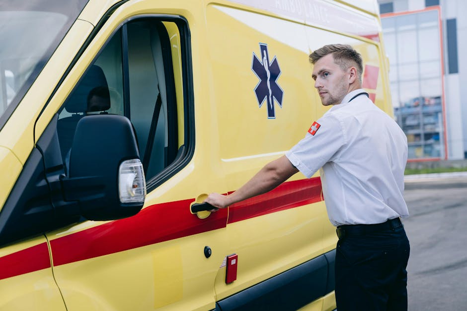 A male ambulance worker with short, light-colored hair, wearing a white medical uniform with a red cross patch on the sleeve, is seen opening the side door of a yellow emergency ambulance vehicle with a red stripe and a blue medical emergency symbol. The scene is outdoors on a paved area, with a modern glass building and some structural elements visible in the background. Inside the ambulance, partially visible, are shelves and compartments likely containing medical equipment. The worker's expression is focused as he prepares the vehicle for a possible rapid response or home relocation involving urgent medical transport, with the loading process implied through his action of opening the door. The vehicle is parked on a roadway or parking area, and the lighting indicates daytime. This scene exemplifies the type of quick-response transportation service that [COMPANY_NAME] could assist with for home removals or emergency logistics related to moving or urgent collections, highlighting the importance of professional vehicle handling in critical situations.