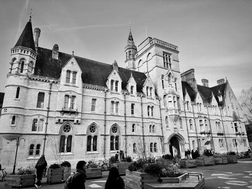 Black and white photograph showing a large historic building with a stone facade, numerous pointed towers, arched windows, and decorative architectural details. The foreground features a paved area with several large wooden planters containing small trees and shrubs, along with parked bicycles. A few pedestrians are walking across the space, some carrying bags or wearing coats, indicating an urban environment. The scene is illuminated by natural daylight, with a partly cloudy sky overhead. This image captures the exterior of a notable structure, likely part of a university or institutional setting, relevant to home relocation, furniture transport, and the logistics involved in moving services that Man with Van Whitton offers, especially around the St Mary's University area.