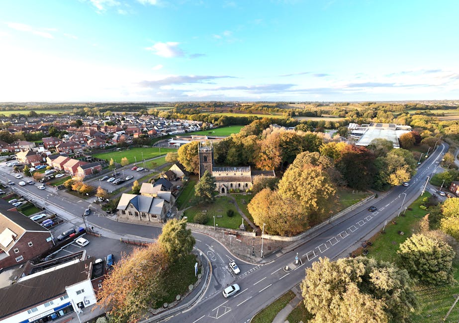 Aerial view of a suburban area featuring a small historic church surrounded by a well-maintained green park with mature trees displaying autumn foliage. Adjacent to the park are residential houses, some with driveways and parked cars. The scene also includes a curved road with several vehicles, pedestrians, and street lamps, capturing the typical surroundings of the St Mary's University area in Whitton. The sky is partly cloudy with sunlight illuminating the scene, highlighting the urban and natural environment. This image reflects the typical setting where house removals, facilitated by companies like Man with Van Whitton, often take place, involving the loading and transport of furniture and boxes across the area for student or home relocations.